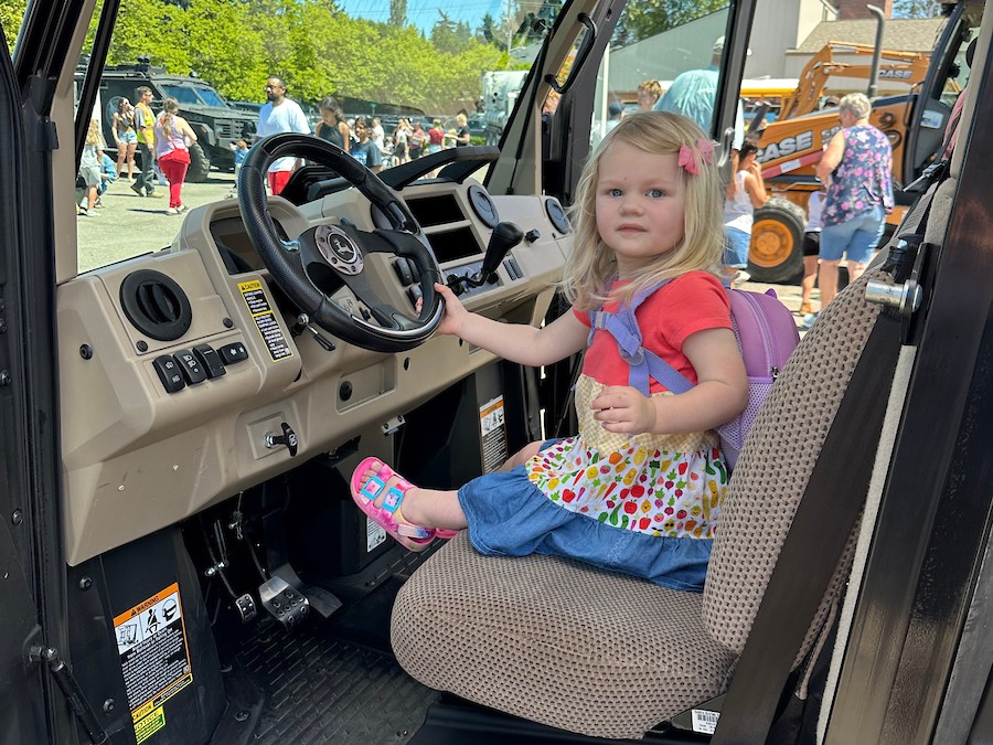 A small girl with blonde hair holds the steering wheel while sitting in the cab of a truck. 
